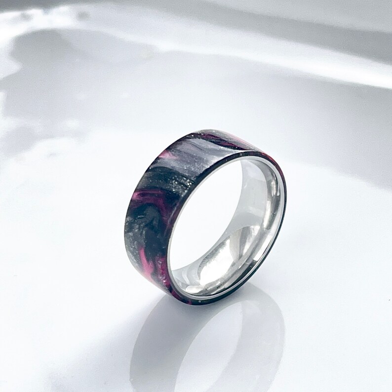 May include: A black and pink resin ring with a silver band. The resin has a marbled pattern.