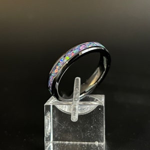 May include: A black tungsten carbide ring with a band of iridescent opal inlay. The ring is on a clear display stand.