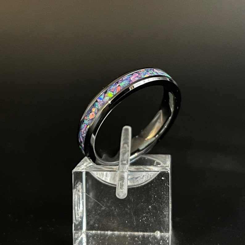 May include: A black tungsten carbide ring with a band of iridescent opal inlay. The ring is on a clear display stand.