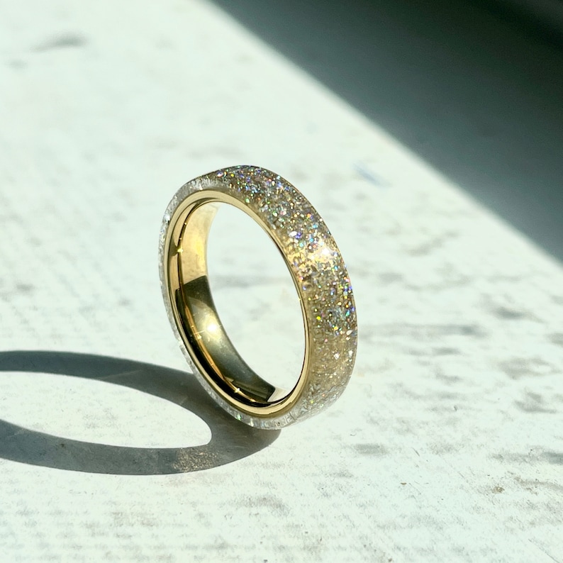 K&ouml;nnte beinhalten: Ein goldfarbener Ring mit einem Band, das mit glitzerndem, glitzer&auml;hnlichem Material bedeckt ist. Der Ring ist vor einem wei&szlig;en Hintergrund platziert, mit einem Schatten, der vom Sonnenlicht geworfen wird. Das Design des Rings ist schlicht, aber elegant.