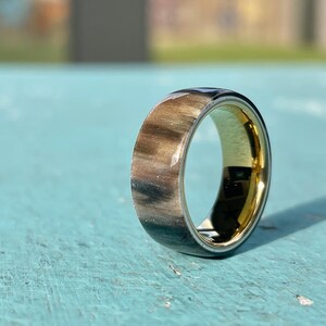 K&ouml;nnte beinhalten: Nahaufnahme eines Rings mit goldener Innenseite und einem Band mit einer Mischung aus Braun-, Schwarz- und Silbert&ouml;nen. Der Ring ist vor einem hellblauen Hintergrund platziert, wobei sich das Sonnenlicht auf der polierten Oberfl&auml;che spiegelt.