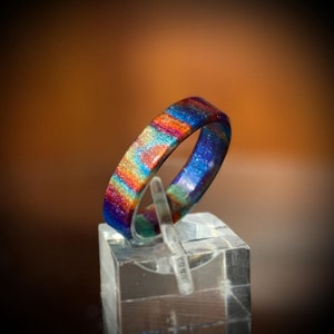 K&ouml;nnte beinhalten: Ein farbenfroher Ring mit Regenbogenmuster, der Farbt&ouml;ne von Blau, Orange, Gelb und Gr&uuml;n aufweist. Der Ring wird auf einem transparenten Acrylst&auml;nder pr&auml;sentiert, der sein glitzerndes Design hervorhebt. Ein einzigartiges Schmuckst&uuml;ck.