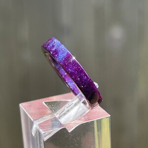 K&ouml;nnte beinhalten: Nahaufnahme eines Rings mit galaxieartigem Design. Der Ring ist haupts&auml;chlich lila mit blauen und wei&szlig;en Streifen und einer glitzernden Textur. Der Ring wird auf einem transparenten, quadratischen St&auml;nder pr&auml;sentiert.