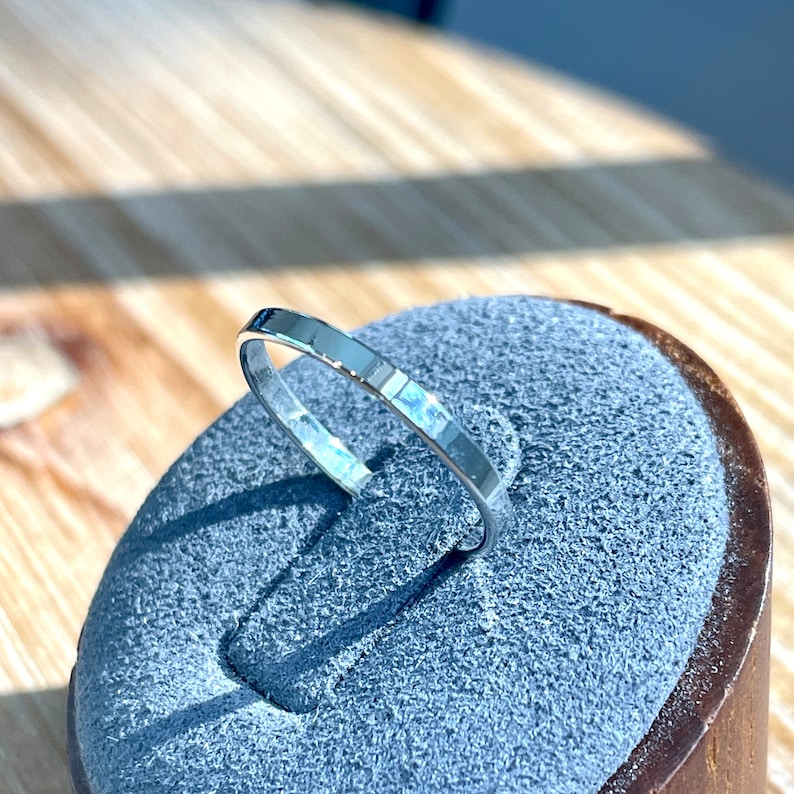 Op de afbeelding: Een zilveren ring met een gehamerde afwerking ligt op een grijze fluwelen ringenstandaard. De ring is simpel en elegant.