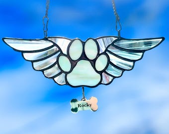 May include: Stained glass pet memorial featuring a paw print with angel wings. The design includes a bone-shaped tag with the name "Rocky". The piece is suspended by a chain, with a blue sky background.