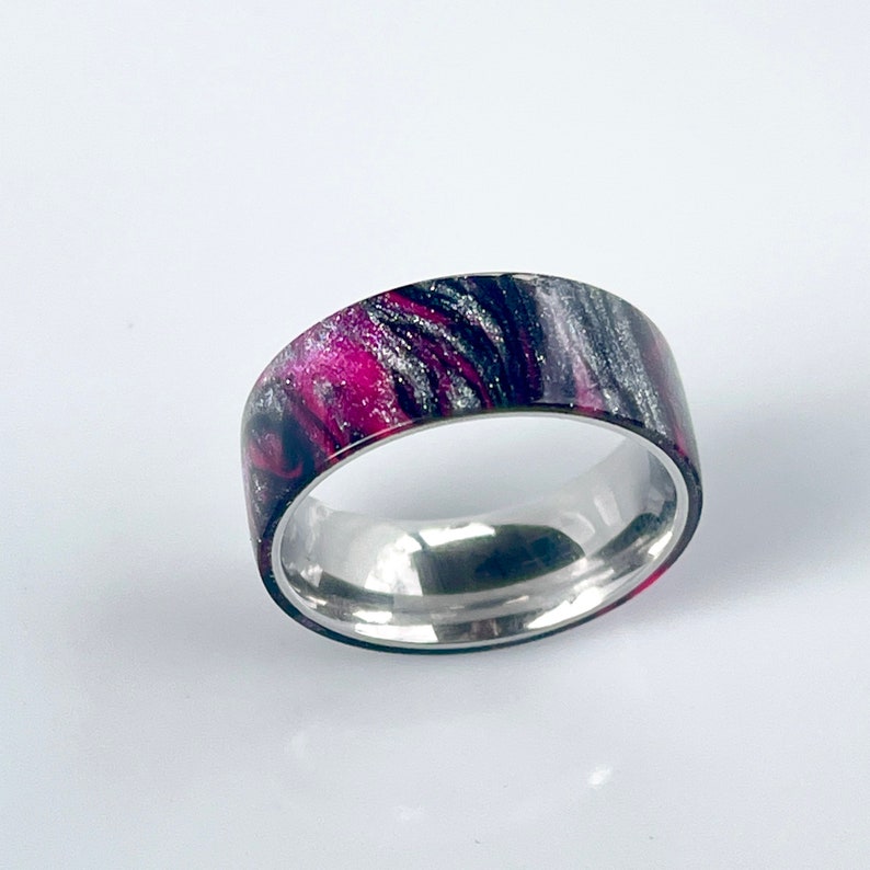 May include: A silver band ring with a black and pink marbled design.