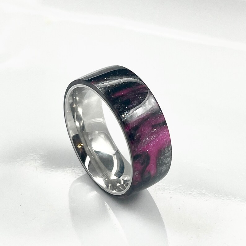 May include: A silver band ring with a black and pink resin inlay. The resin has a swirling pattern with white glitter embedded in it.