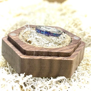 May include: A silver ring with a purple and blue inlay sits in a wooden ring box. The box is hexagonal and has a dark brown finish.