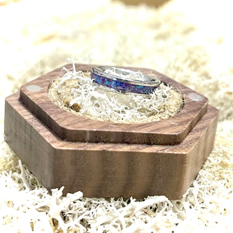 May include: A silver ring with a purple and blue inlay sits in a wooden ring box. The box is hexagonal and has a dark brown finish.