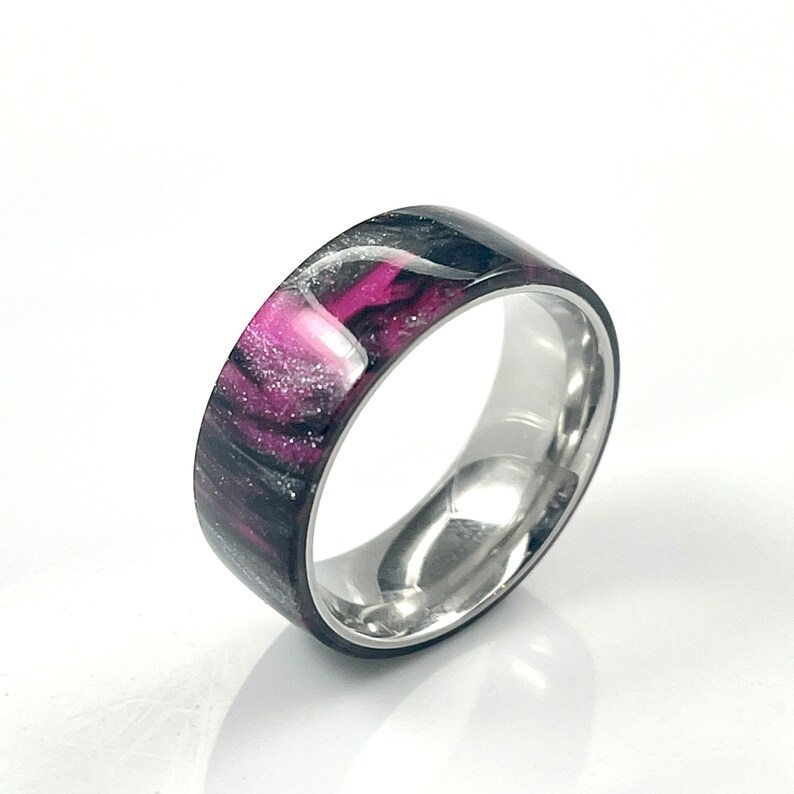 May include: A black and silver ring with a pink and white marbled design. The ring is made of stainless steel and has a polished finish.
