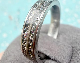 May include: A silver ring with a clear band filled with sparkling, iridescent glitter. The ring has a polished, metallic finish and is set against a soft gray background. The ring's design is modern and eye-catching.