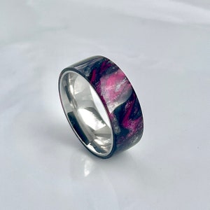 May include: A silver band ring with a black and pink resin inlay. The resin has a swirling pattern with silver glitter.
