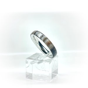 K&ouml;nnte beinhalten: Ein silberner Ring mit einem Band mit einem gesprenkelten Muster aus Wei&szlig;, Braun und Grau. Der Ring wird auf einem transparenten, quadratischen St&auml;nder vor wei&szlig;em Hintergrund pr&auml;sentiert. Das Design des Rings ist schlicht und elegant.