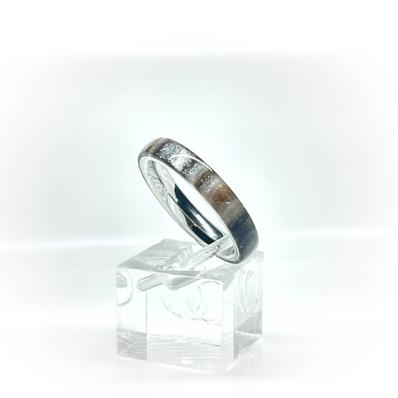 K&ouml;nnte beinhalten: Ein silberner Ring mit einem Band mit einem gesprenkelten Muster aus Wei&szlig;, Braun und Grau. Der Ring wird auf einem transparenten, quadratischen St&auml;nder vor wei&szlig;em Hintergrund pr&auml;sentiert. Das Design des Rings ist schlicht und elegant.