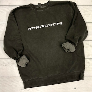 May include: A dark green sweatshirt with a white print of the coordinates "30°12'56.0"N 92°02'12.7"W"