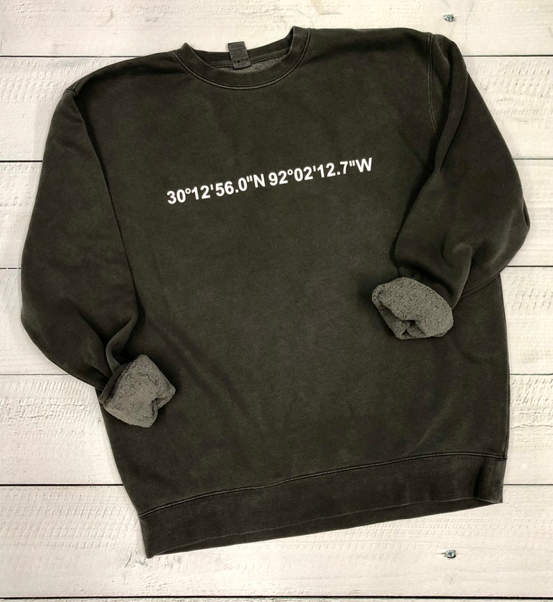 Puede incluir: Una sudadera de color verde oscuro con una impresi&oacute;n blanca de las coordenadas "30&deg;12'56.0"N 92&deg;02'12.7"W"