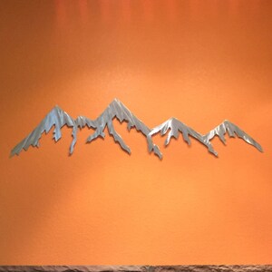 K&ouml;nnte beinhalten: Ein silberfarbenes Wandkunstobjekt aus Metall in Form eines Gebirgszuges mit gezackten, spitzen Gipfeln. Die Skulptur ist an einer einfarbigen orangefarbenen Wand &uuml;ber einem Steinkamin mit schwarzer Glast&uuml;r und einer h&ouml;lzernen Schrankt&uuml;r angebracht.