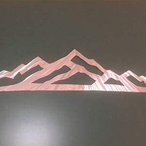 May include: A rose gold metal wall art piece featuring a stylized mountain range silhouette. The design consists of a continuous, thin, angular outline of peaks and valleys with a metallic, reflective finish and a flat, horizontal base.