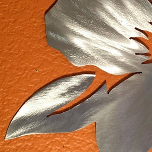 Puede incluir: Una escultura de pared de metal plateado en forma de flor con un acabado cepillado. La flor tiene un p&eacute;talo grande y un p&eacute;talo m&aacute;s peque&ntilde;o. La escultura est&aacute; montada en una pared naranja texturizada.