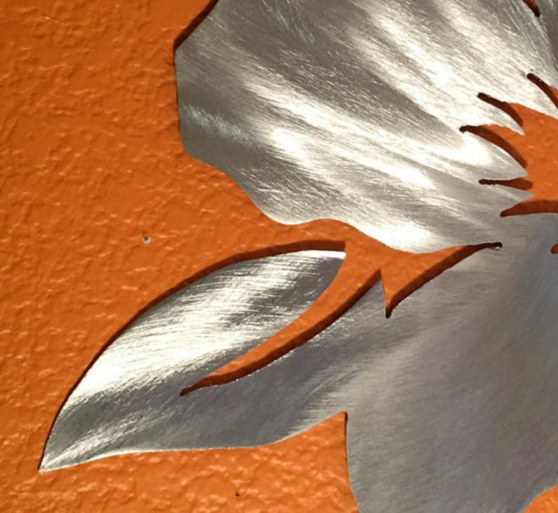 Puede incluir: Una escultura de pared de metal plateado en forma de flor con un acabado cepillado. La flor tiene un p&eacute;talo grande y un p&eacute;talo m&aacute;s peque&ntilde;o. La escultura est&aacute; montada en una pared naranja texturizada.