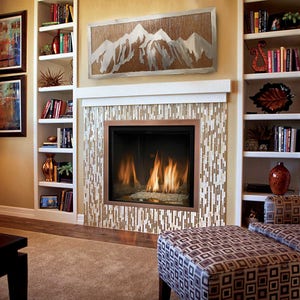 May include: A cozy living room with a fireplace, bookshelves, and artwork. The fireplace has a mosaic tile surround and a copper-colored frame. A mountain-themed artwork hangs above the fireplace. The room is furnished with a patterned armchair and ottoman.