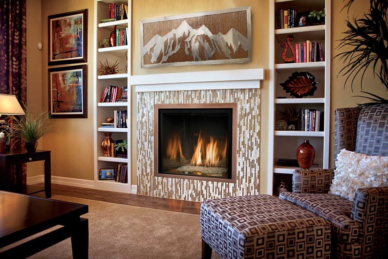 May include: A cozy living room with a fireplace, bookshelves, and artwork. The fireplace has a mosaic tile surround and a copper-colored frame. A mountain-themed artwork hangs above the fireplace. The room is furnished with a patterned armchair and ottoman.