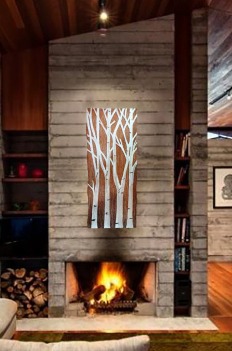 May include: A rectangular wooden artwork featuring a design of white birch trees against a brown background. The artwork is mounted on a gray stone fireplace, with a fire burning in the hearth below. The room has a modern, rustic aesthetic.