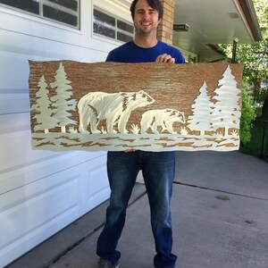 K&ouml;nnte beinhalten: Metall-Wandkunst mit einem B&auml;ren und einem Jungtier in einer Waldszene. Das Kunstwerk hat einen strukturierten braunen Hintergrund mit wei&szlig;en B&auml;ren- und Baum-Silhouetten. Das rechteckige St&uuml;ck ist etwa 100 cm breit. Ein dekorativer Artikel f&uuml;r das Zuhause.