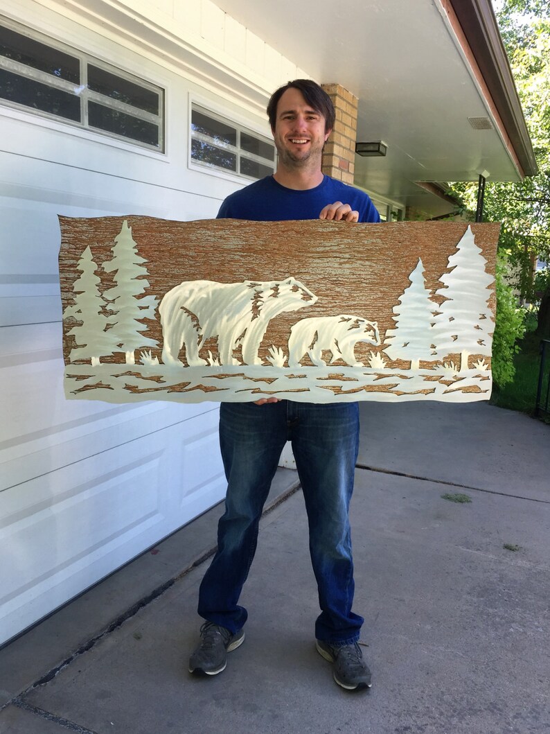 K&ouml;nnte beinhalten: Metall-Wandkunst mit einem B&auml;ren und einem Jungtier in einer Waldszene. Das Kunstwerk hat einen strukturierten braunen Hintergrund mit wei&szlig;en B&auml;ren- und Baum-Silhouetten. Das rechteckige St&uuml;ck ist etwa 100 cm breit. Ein dekorativer Artikel f&uuml;r das Zuhause.