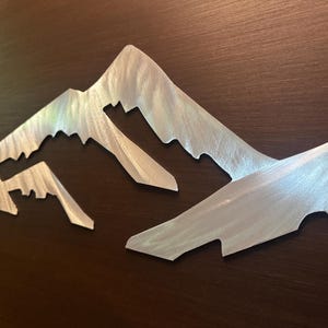 May include: A silver-colored metal wall art piece featuring a stylized mountain range silhouette. The surface of the metal has a brushed texture with visible directional grain, creating a reflective finish across the jagged peaks and slopes.