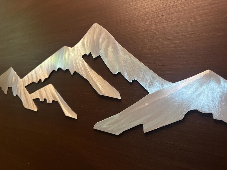 May include: A silver-colored metal wall art piece featuring a stylized mountain range silhouette. The surface of the metal has a brushed texture with visible directional grain, creating a reflective finish across the jagged peaks and slopes.
