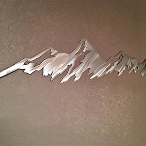 May include: A metal mountain range wall art piece. The artwork is cut from a silver-colored metal and depicts a jagged mountain range silhouette. The piece is mounted on a textured, neutral-toned wall, creating a contrast between the smooth metal and the wall's surface.