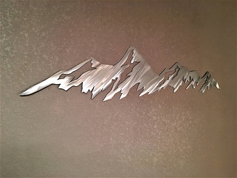 May include: A metal mountain range wall art piece. The artwork is cut from a silver-colored metal and depicts a jagged mountain range silhouette. The piece is mounted on a textured, neutral-toned wall, creating a contrast between the smooth metal and the wall's surface.