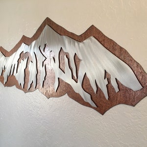 May include: A wall-mounted metal art piece featuring a layered design with a silver-toned mountain range silhouette positioned over a textured, copper-colored backing plate. The mountain peaks have a brushed finish with vertical lines.