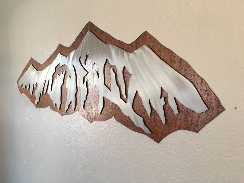 May include: A wall-mounted metal art piece featuring a layered design with a silver-toned mountain range silhouette positioned over a textured, copper-colored backing plate. The mountain peaks have a brushed finish with vertical lines.