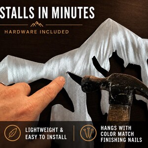 May include: A silver-colored metal mountain range wall art piece featuring a brushed finish. The text reads: INSTALLS IN MINUTES, HARDWARE INCLUDED, LIGHTWEIGHT & EASY TO INSTALL, and HANGS WITH COLOR MATCH FINISHING NAILS. A hammer is shown.