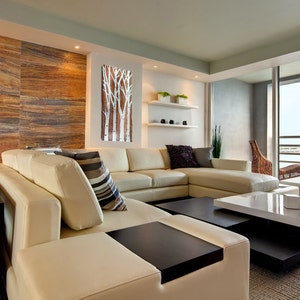 May include: A modern living room featuring a large cream-colored sectional sofa with a dark wood coffee table. A decorative wall art piece with white birch tree silhouettes hangs on the wall. The room has a neutral color palette with wood and stone accents.