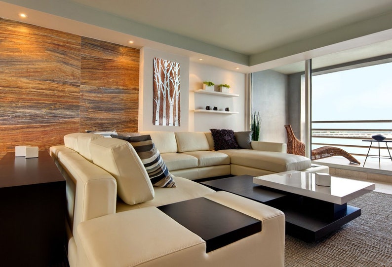 May include: A modern living room featuring a large cream-colored sectional sofa with a dark wood coffee table. A decorative wall art piece with white birch tree silhouettes hangs on the wall. The room has a neutral color palette with wood and stone accents.