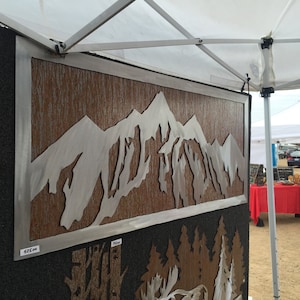 May include: Metal wall art featuring a mountain range silhouette. The artwork has a silver-colored mountain design set against a textured brown background, framed in silver. The piece is displayed outdoors under a white canopy.