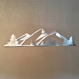 May include: A metal wall art sculpture depicting a mountain range with trees in front. The sculpture is silver in color and has a brushed finish.