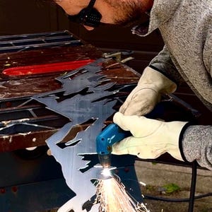 May include: A person wearing protective eyewear and beige leather gloves uses a blue plasma cutter to carve a jagged mountain range design into a flat sheet of dark gray metal, creating bright orange sparks at the point of contact.