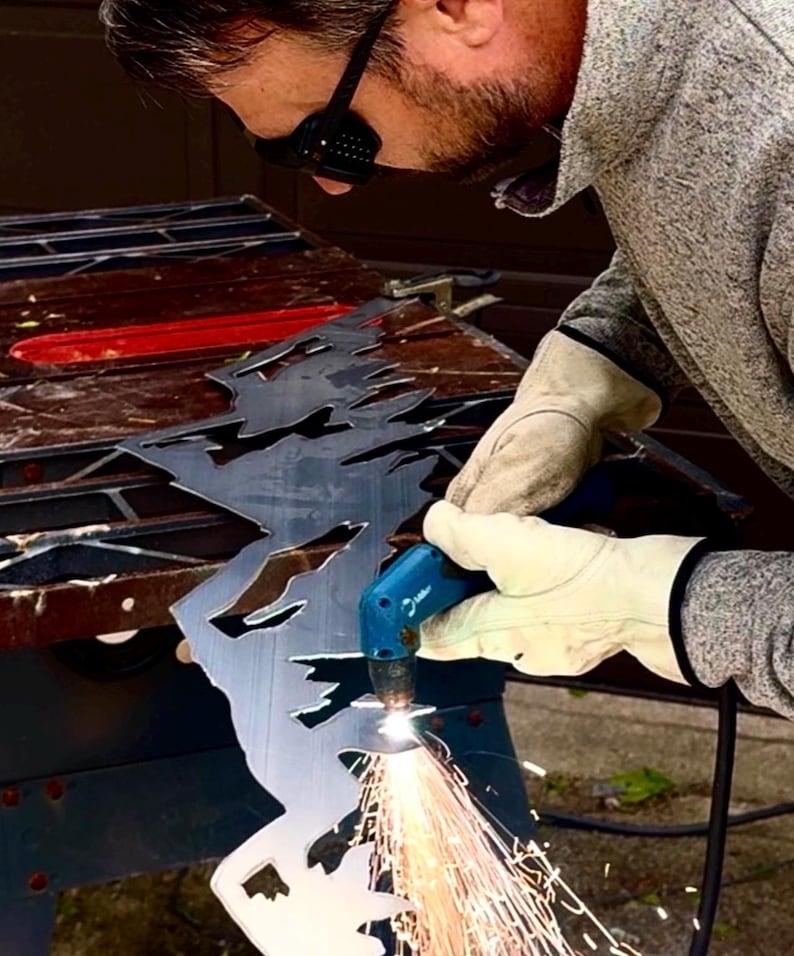 May include: A person wearing protective eyewear and beige leather gloves uses a blue plasma cutter to carve a jagged mountain range design into a flat sheet of dark gray metal, creating bright orange sparks at the point of contact.