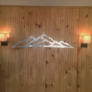 May include: A silver-colored metal wall art piece featuring a stylized mountain range silhouette with jagged peaks and valleys. The flat, reflective metal cutout is mounted horizontally, showcasing a clean, minimalist outline of a mountain landscape.