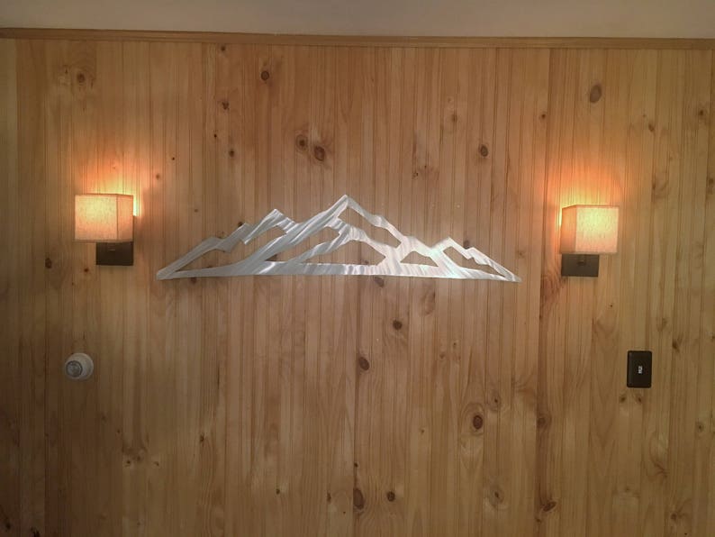 May include: A silver-colored metal wall art piece featuring a stylized mountain range silhouette with jagged peaks and valleys. The flat, reflective metal cutout is mounted horizontally, showcasing a clean, minimalist outline of a mountain landscape.