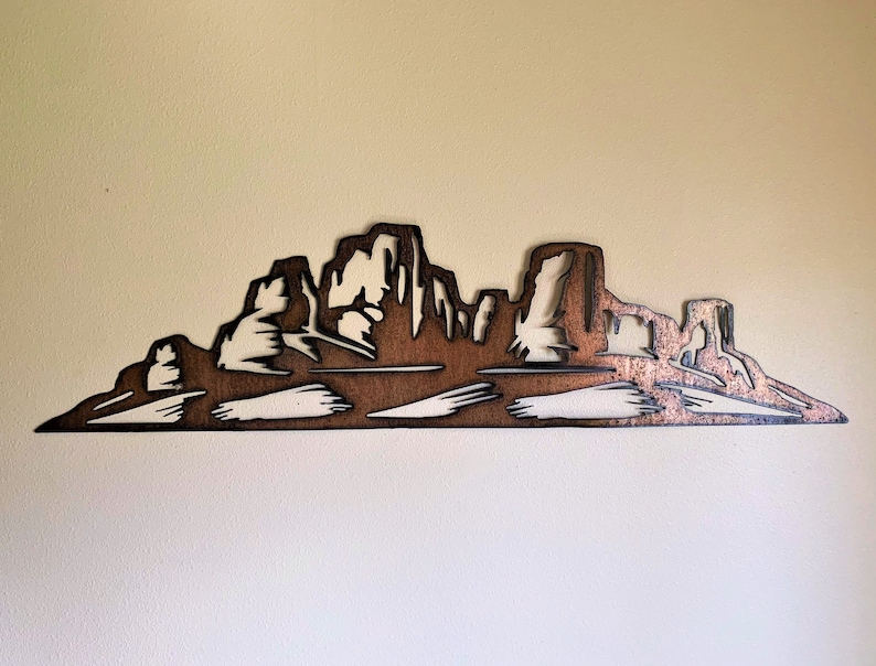 Puede incluir: Una pieza de arte mural de metal marr&oacute;n que representa una silueta de cordillera. La obra de arte presenta un dise&ntilde;o recortado detallado con picos y valles, y una orientaci&oacute;n horizontal. La pieza est&aacute; montada en una pared de color claro, creando un contraste.
