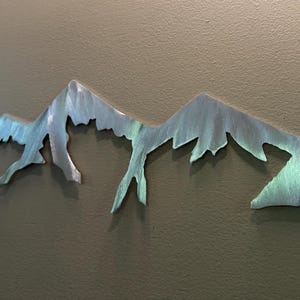 K&ouml;nnte beinhalten: Ein horizontales Wandkunstobjekt aus Metall, das die Silhouette eines Gebirgszuges darstellt. Die Oberfl&auml;che hat ein geb&uuml;rstetes Silber-Finish mit subtilen irisierenden Blau- und Gr&uuml;nt&ouml;nen, die gezackte Gipfel und tiefe T&auml;ler zeigen.