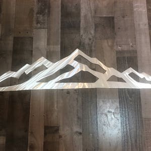 May include: A wide, horizontal wall art piece crafted from brushed silver metal, featuring a continuous, stylized mountain range silhouette with sharp, angular peaks and valleys, showcasing a reflective, metallic finish across the entire surface.