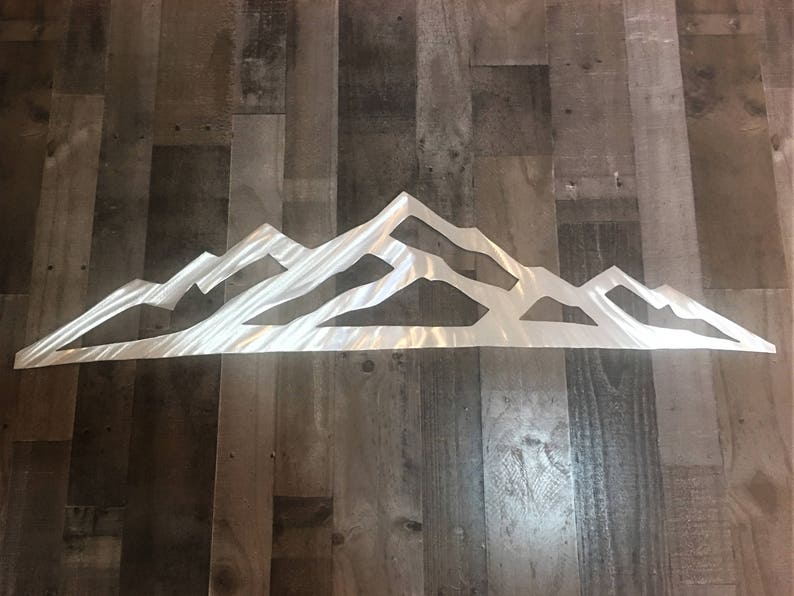 May include: A wide, horizontal wall art piece crafted from brushed silver metal, featuring a continuous, stylized mountain range silhouette with sharp, angular peaks and valleys, showcasing a reflective, metallic finish across the entire surface.