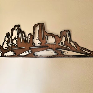 Puede incluir: Una pieza de arte mural de metal que representa un paisaje des&eacute;rtico con monta&ntilde;as y mesas. La obra de arte est&aacute; cortada de metal marr&oacute;n, con espacio negativo que crea las formas de las formaciones. La pieza est&aacute; montada en una pared de color neutro.