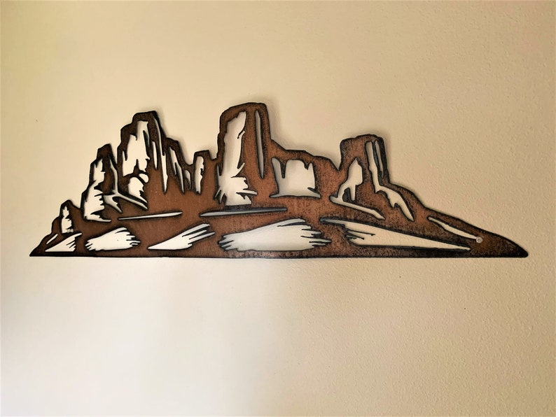 Puede incluir: Una pieza de arte mural de metal que representa un paisaje des&eacute;rtico con monta&ntilde;as y mesas. La obra de arte est&aacute; cortada de metal marr&oacute;n, con espacio negativo que crea las formas de las formaciones. La pieza est&aacute; montada en una pared de color neutro.
